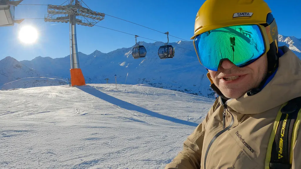 A skier in a helmet and mirrored goggles on the slope, with a gondola and snowy peaks in the background